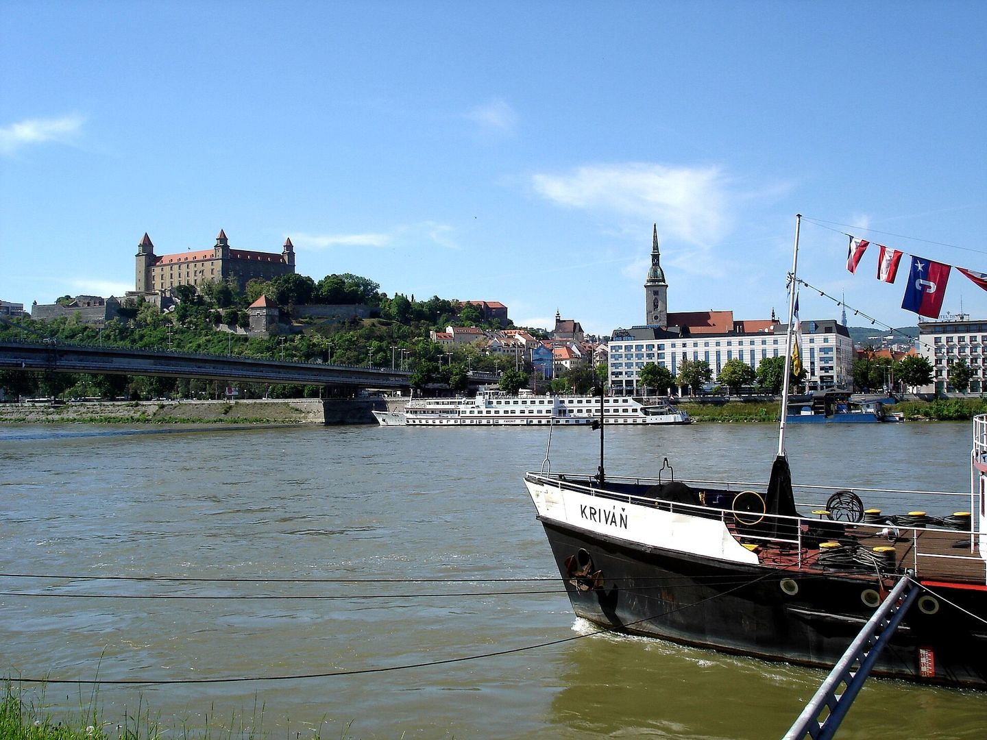 Panoramic view of the Danube River in Bratislava with cargo and passenger boats moored along the embankment and Bratislava Castle's white walls and towers rising on the hill behind the Old Town