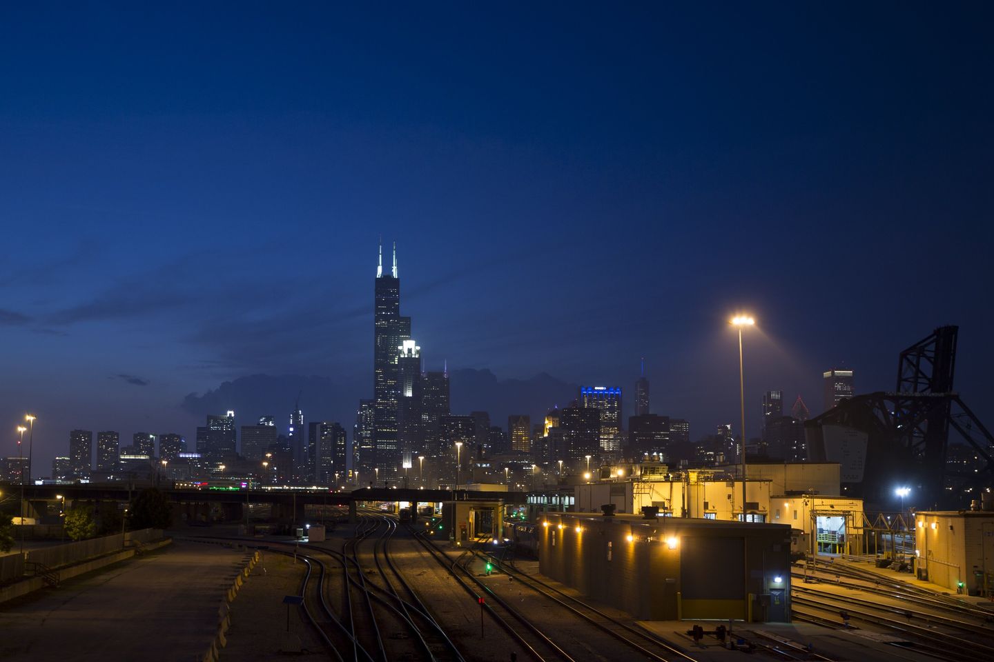 The Chicago skyline lit at dusk, with the black steel of the Willis Tower and the cluster of Loop skyscrapers rising above the lakefront under a deep blue evening sky