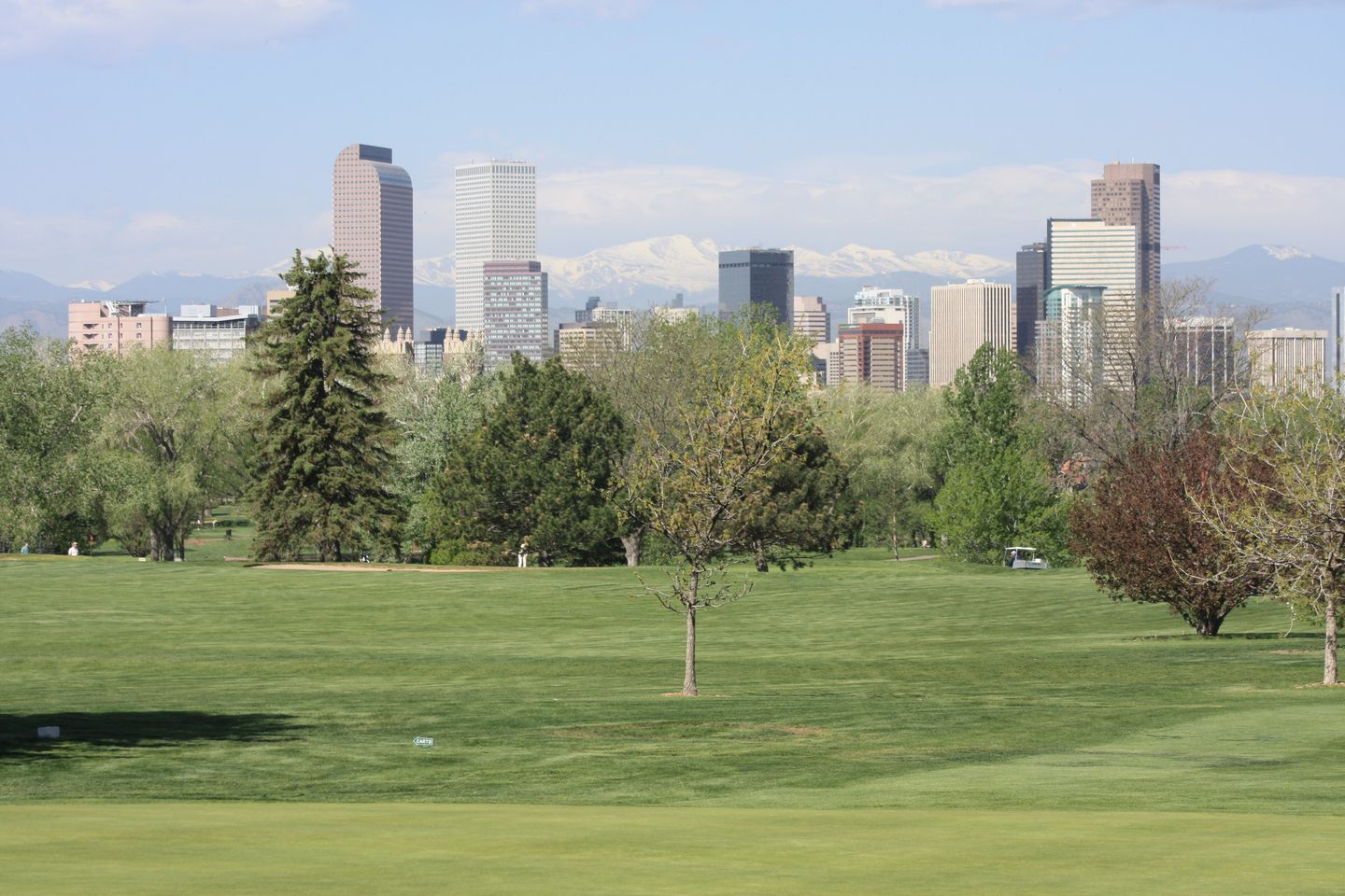 The downtown Denver skyline of glass towers seen from City Park Golf Course, with the snow-dusted peaks of the Front Range and Mount Evans rising behind it