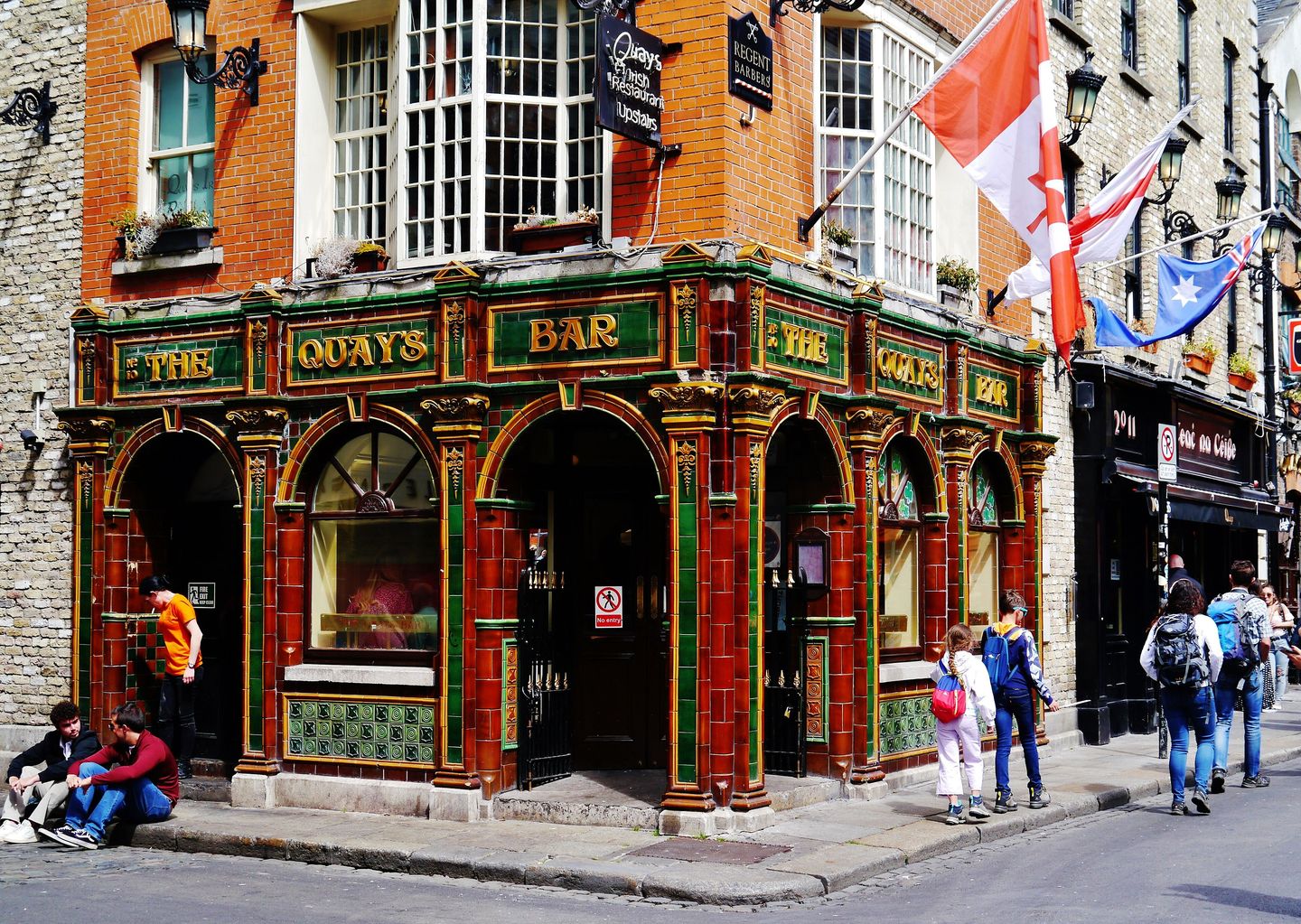 The ornate red, green and gold Victorian tiled corner facade of The Quay's Bar in Temple Bar, Dublin, with national flags hanging above the arched windows and pedestrians on the cobbled street below