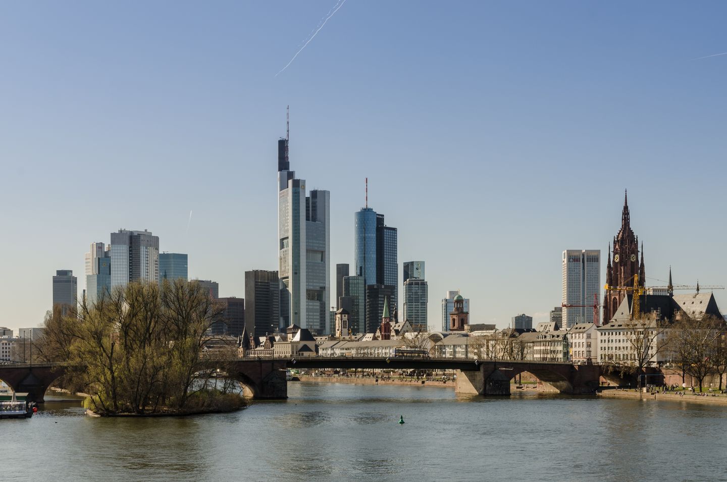 The Frankfurt skyline of glass office towers reflected in the Main River at dusk, seen from the riverbank with the old town rooftops visible at the water's edge
