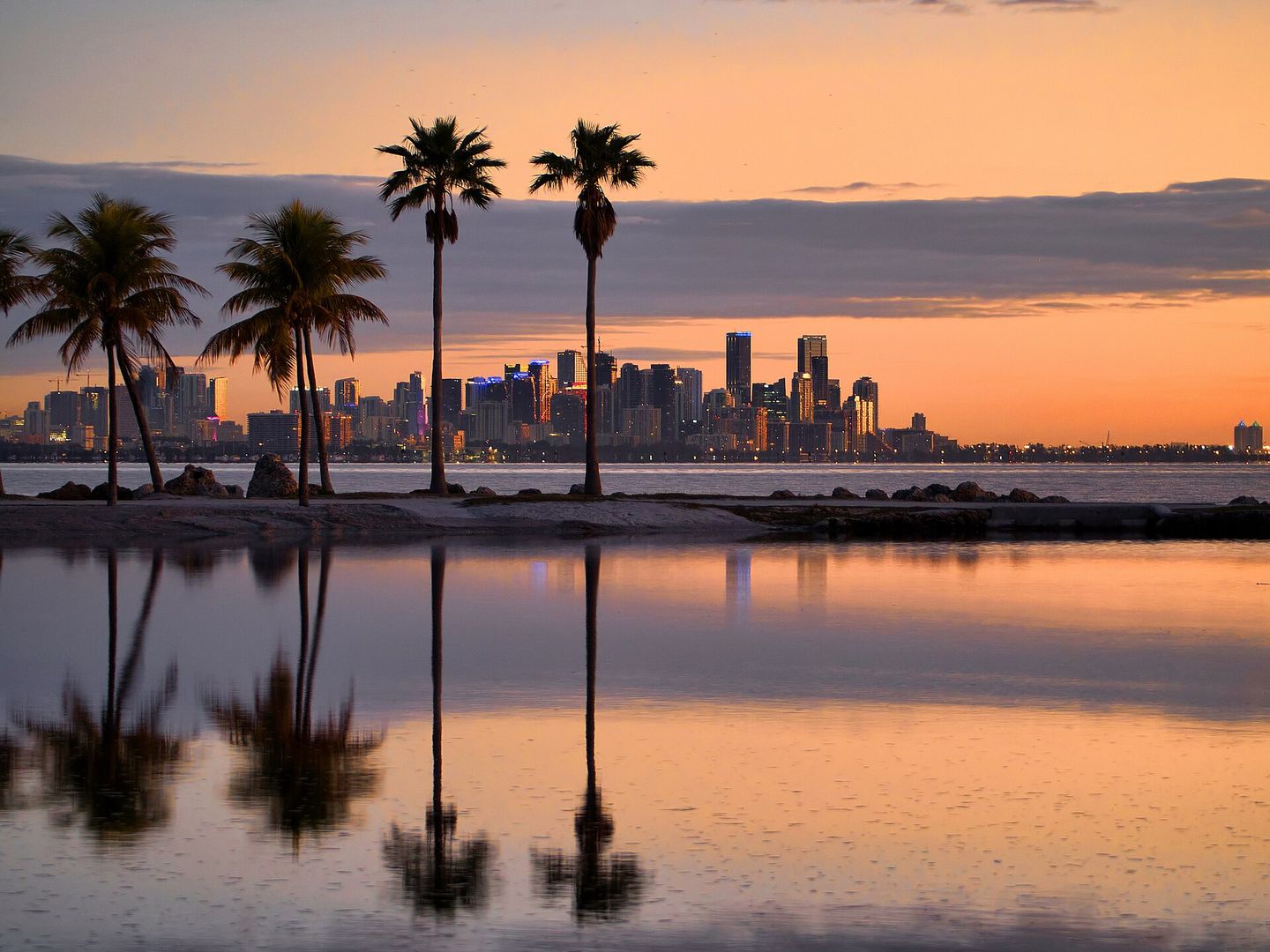Silhouetted palm trees on a spit of sand at Matheson Hammock Park at dawn, with the lit Miami downtown skyline reflected in the still water of Biscayne Bay under an orange sky