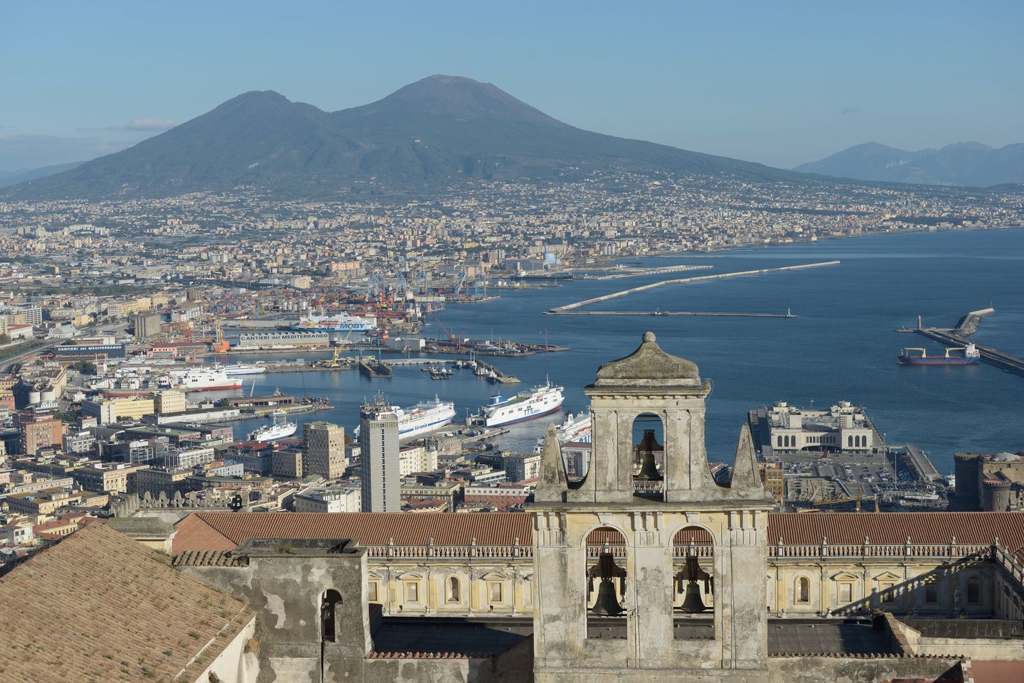 The rooftops of Naples' old town seen from Castel Sant'Elmo, with the dome of the Certosa di San Martino in the foreground, the harbour beyond, and Mount Vesuvius rising across the bay
