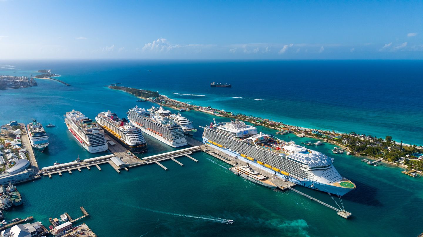 Aerial view of Nassau Cruise Port with six large cruise ships moored side by side along the wharf, the low green shoreline of Paradise Island curving away to the right, and turquoise shallows giving way to open Atlantic blue beyond