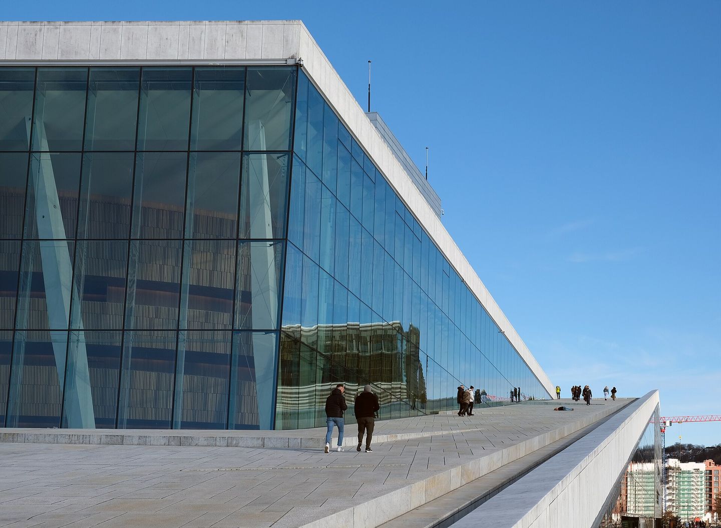 The white sloping marble roof of the Oslo Opera House rising out of the fjord at Bjørvika, with people walking up the incline and the harbour behind