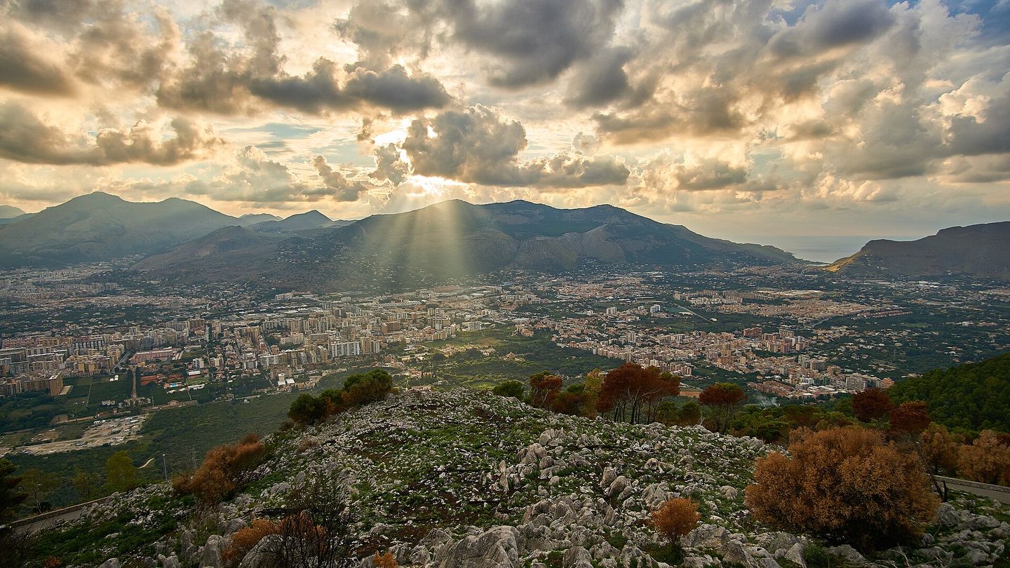 Wide sunset view of Palermo's rooftops and the Bay of Palermo seen from Monte Pellegrino, with the city spreading out toward the sea under a clouded orange sky