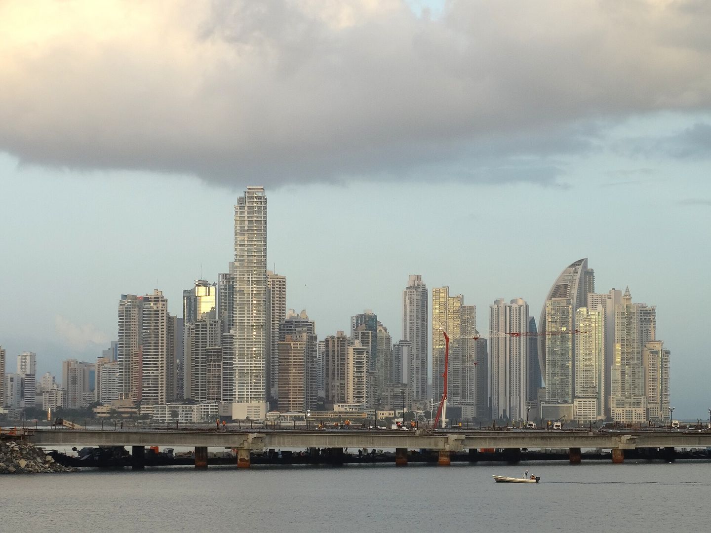 The high-rise skyline of Panama City seen at dusk across the bay from Casco Viejo, a curved sail-shaped tower and construction cranes among the apartment blocks, with a low causeway and a small boat in the foreground under heavy cloud