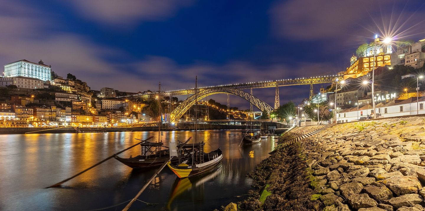The Dom Luís I iron bridge spanning the Douro River at dusk, linking Porto's Ribeira district to the port wine cellars of Vila Nova de Gaia, with the hillside houses of both banks lit against a deep blue sky