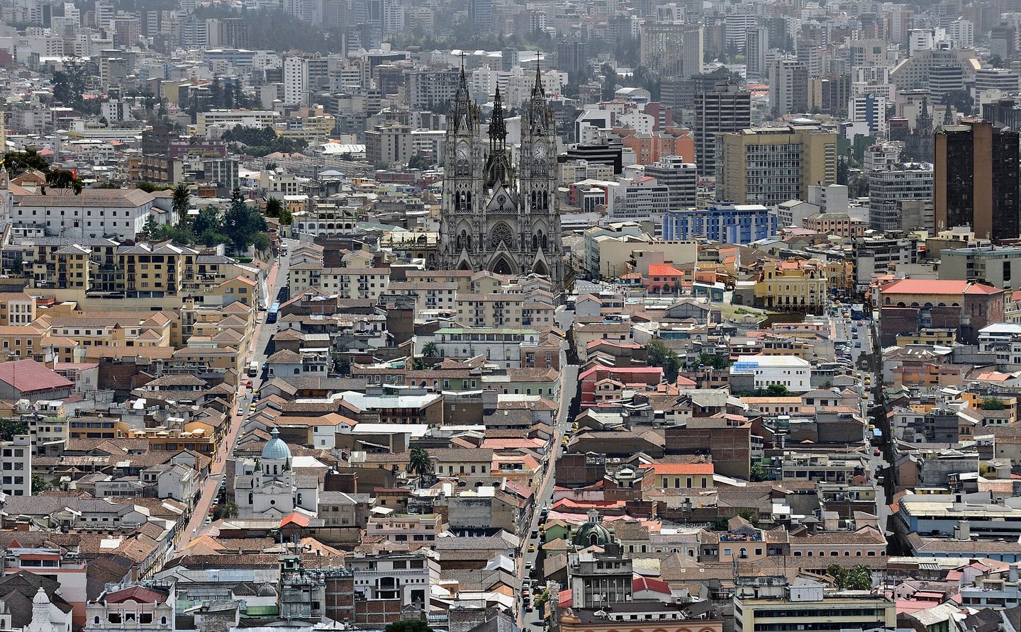 Aerial view over the red-tiled roofs of Quito's historic centre from El Panecillo hill, with the Gothic spires of the Basílica del Voto Nacional rising above the colonial rooftops and the modern city stretching into haze beyond