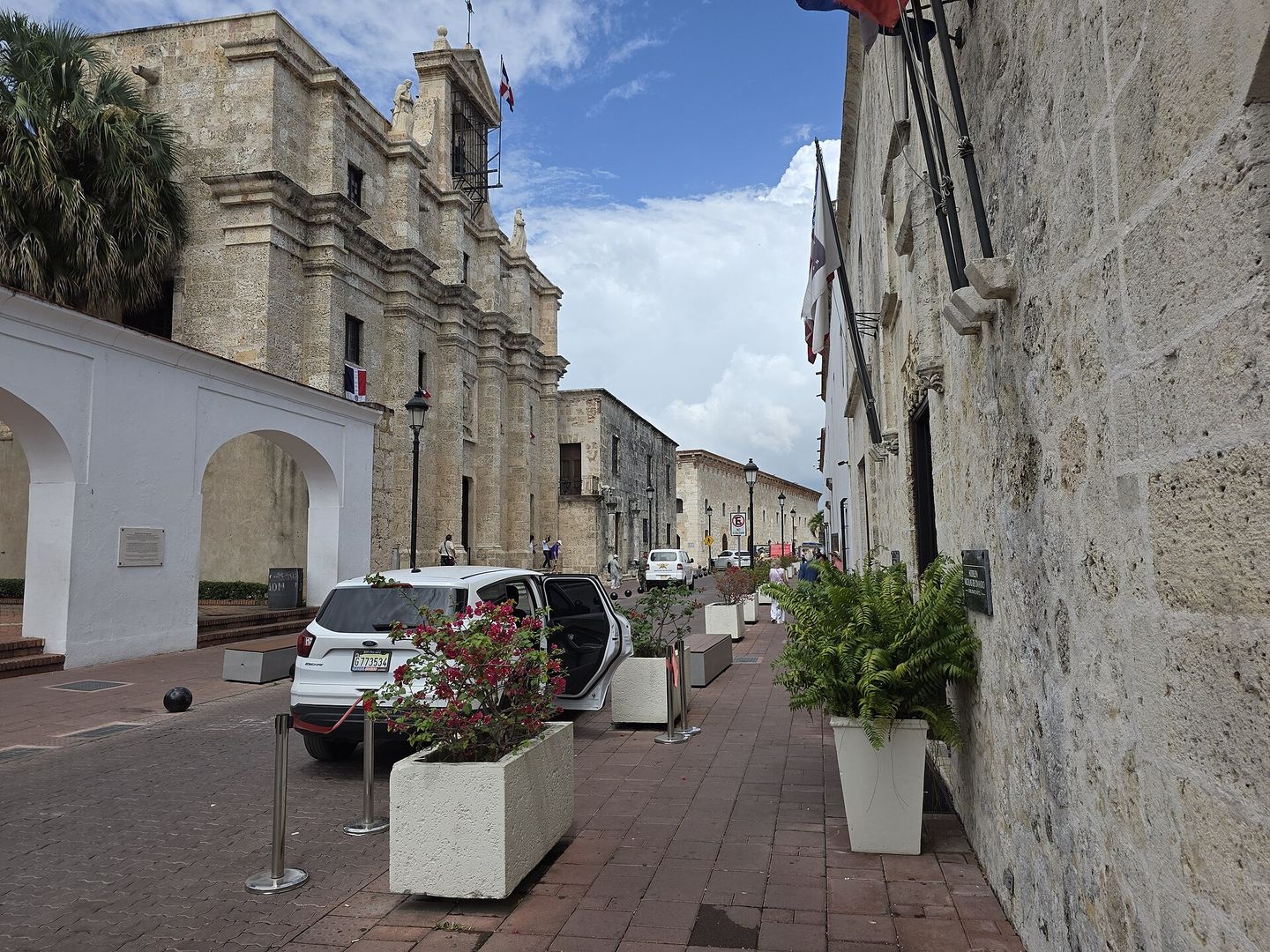 Calle Las Damas in Santo Domingo's Zona Colonial, with the limestone baroque facade and bell tower of the Panteón Nacional flying the Dominican flag on the left, white planters of bougainvillea along the brick-paved pavement, and colonial stone walls receding down the street