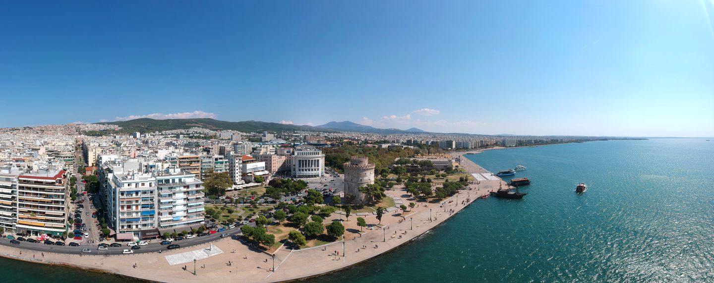 Aerial view from the sea of Thessaloniki's dense waterfront skyline and promenade curving along the Thermaic Gulf, with the city's hills rising behind