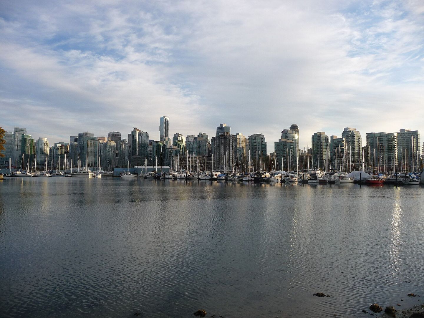 Downtown Vancouver's glass towers reflected in the still water of Coal Harbour marina, seen across a row of moored sailboats from the Stanley Park seawall on a bright afternoon
