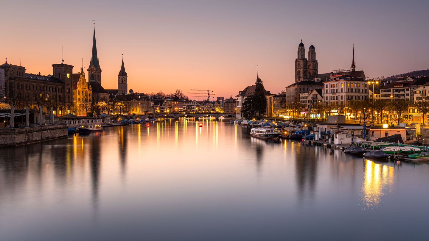 The medieval spires of Zurich's Grossmünster and old town reflected in the Limmat river at dusk, seen from the Quaibrücke